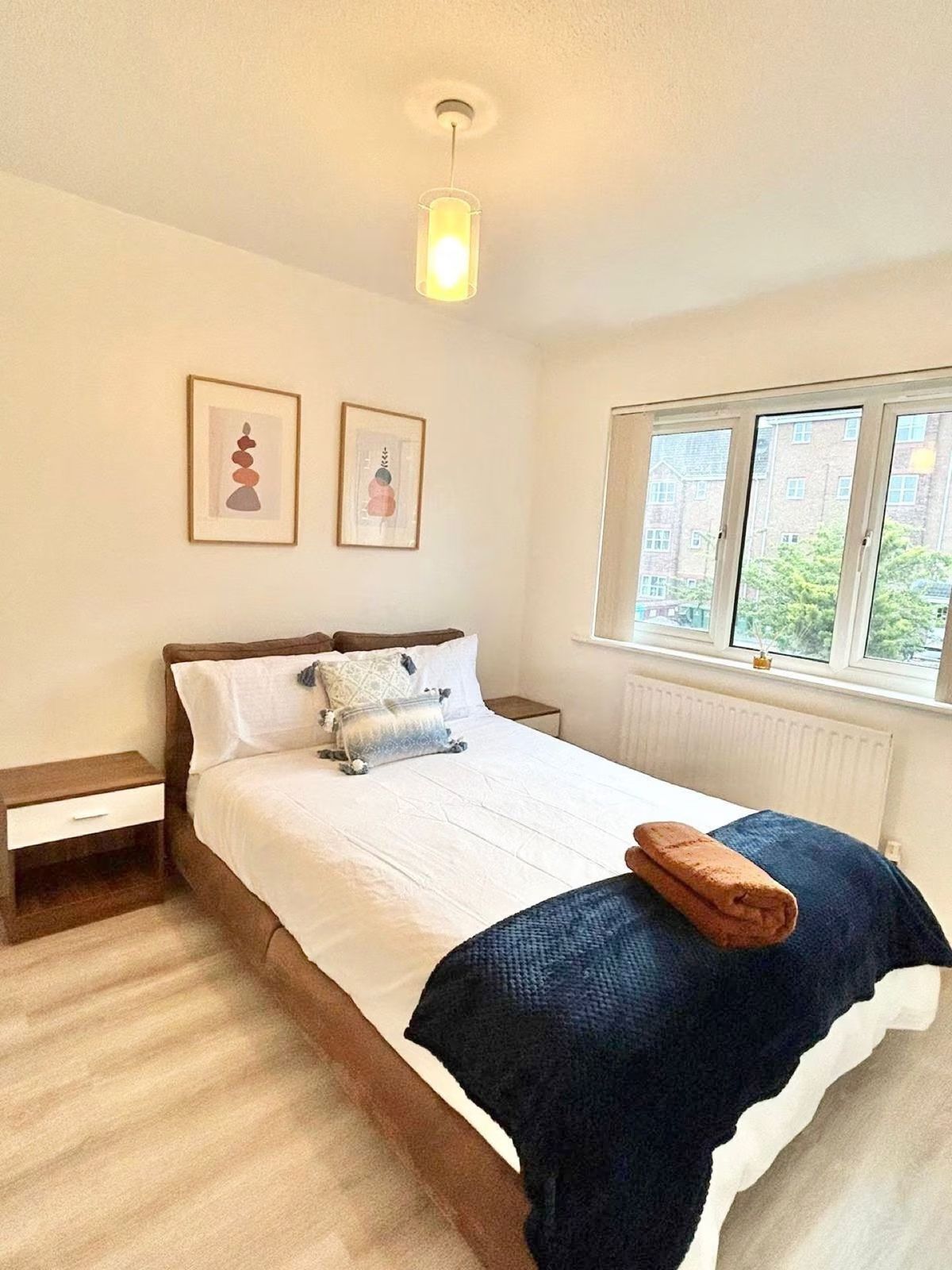 Master bedroom — double bed with white linens and navy throw, two bedside drawers, art prints, ensuite access
