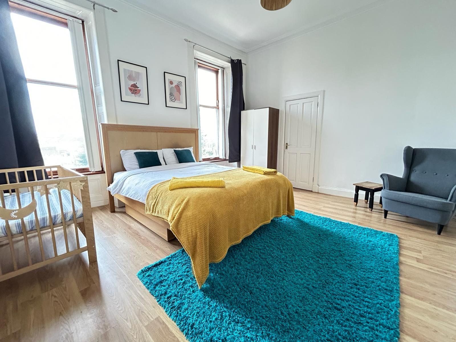 Master bedroom — king bed with mustard blanket and teal accents, cot for the little one, blue armchair, two tall sash windows